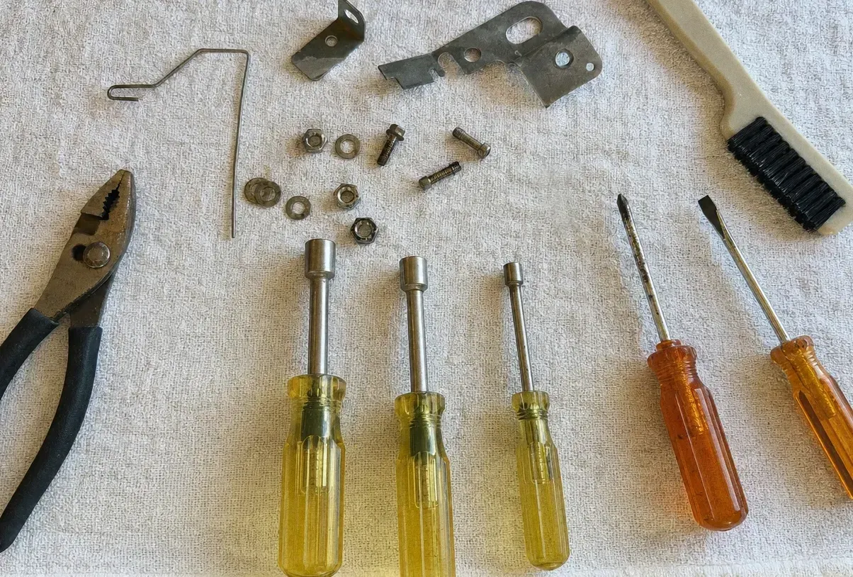 Overhead view of a bent wire clip, a bracket and a scatter of hex nuts and washers beside four screwdrivers, pliers and a brush
